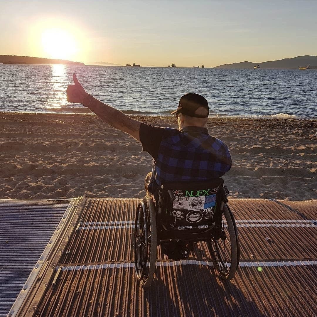 Kevin on a beach matting at sunset, facing the water in his wheelchair with one arm raised