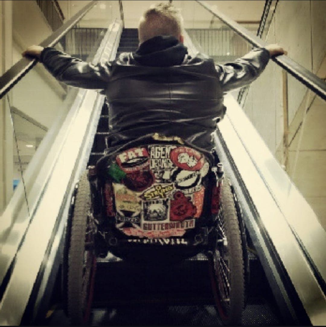 Kevin from behind riding an escalator in his wheelchair, the back of the chair covered in punk band stickers