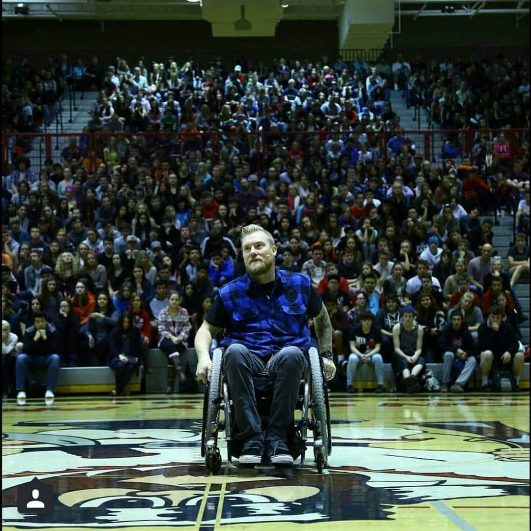 Kevin speaking from his wheelchair on a gymnasium floor, a packed crowd of students in the stands behind him