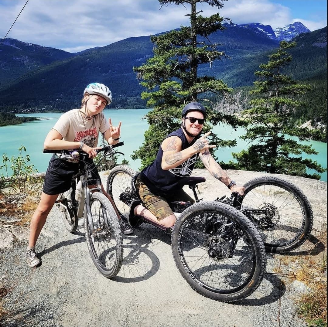 Kevin on his handcycle beside a friend on a mountain bike, a turquoise lake and mountains behind them