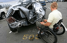 Kevin in his wheelchair, facing the crushed wreck of his car in an impound lot