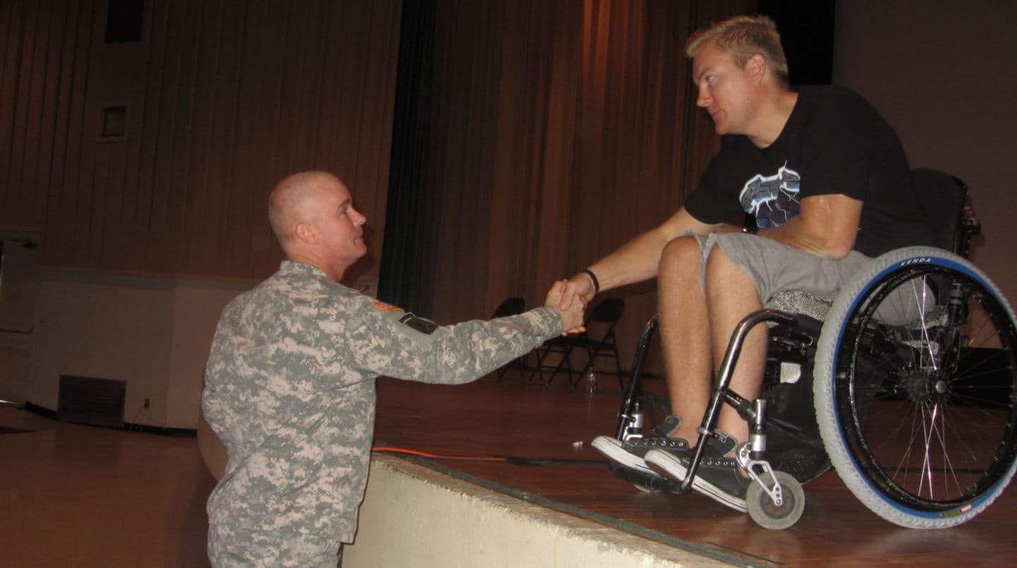 A US Army soldier kneeling to shake Kevin's hand at the edge of a stage after a presentation