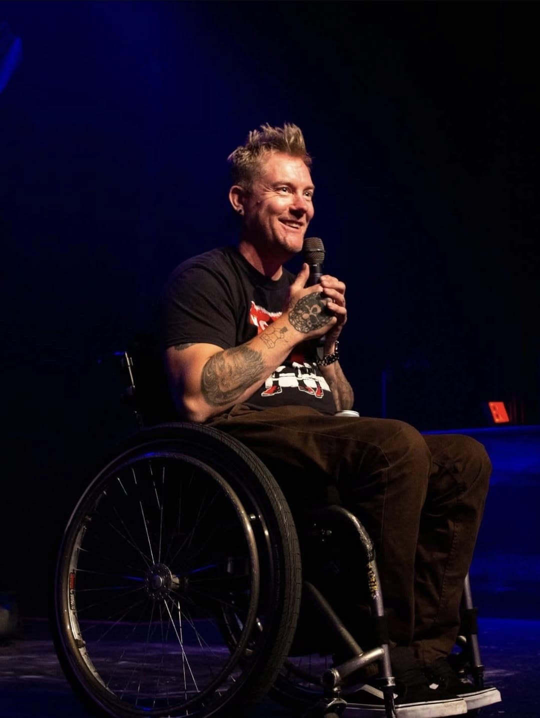 Kevin Brooks on stage in his wheelchair at the Rickshaw in Vancouver, microphone in one tattooed hand, grinning out at the room under a blue stage wash