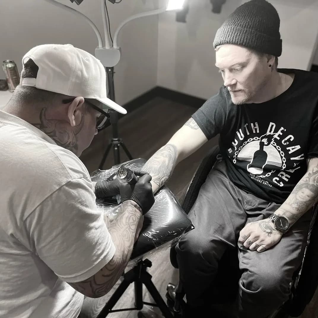 Black and white photograph of Kevin having a tattoo done, the artist working on his arm.